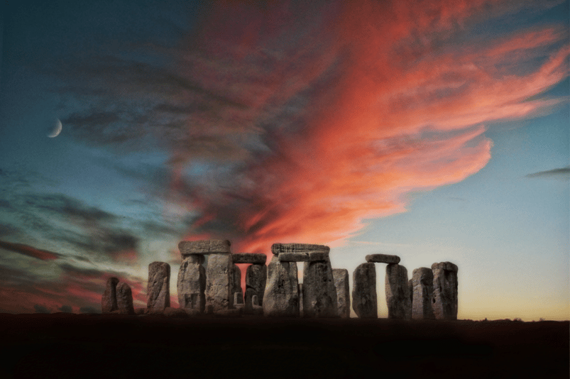 Une photo de Stonehenge au crépuscule ; les nuages ​​au-dessus sont teintés de rouge et sur la gauche un croissant de lune est visible.